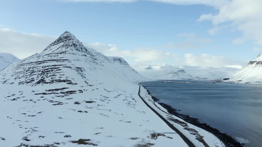 Road parallels shore at base of snow covered mountain on a cloudy day