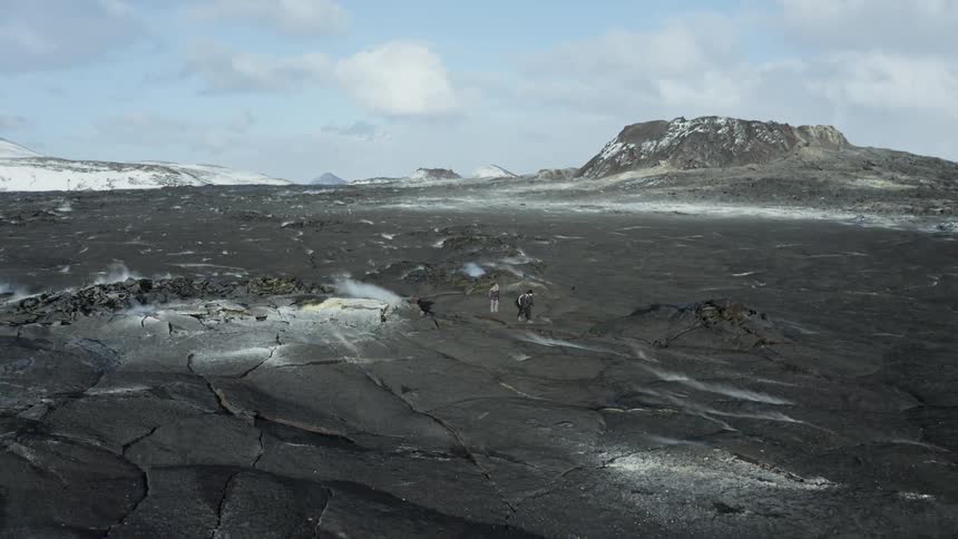 Two people hiking on volcanic rocks with erosion and snow around them on a blue sky day with clouds