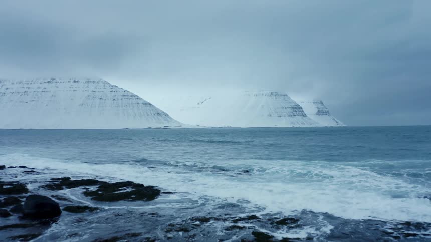 A coast of a sea and there are two huge mountains covered in snow in the background