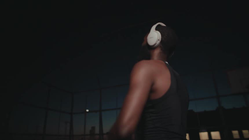 In a fenced basketball court at night a man throws up a basketball and catches it as it bounces