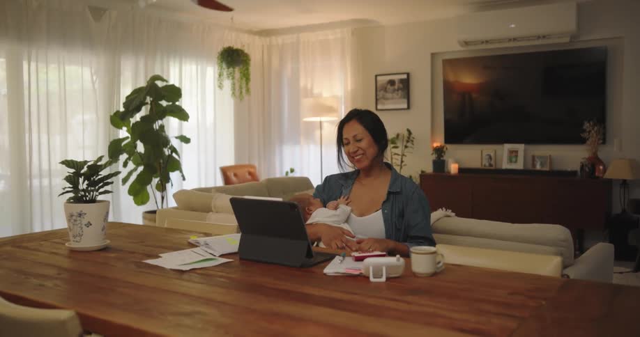 A seated mother is breastfeeding her child while looking and smiling at her laptop screen