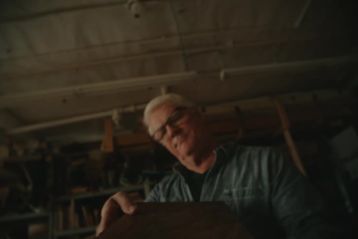 Older male woodworker picks up wooden object and carries it through workshop