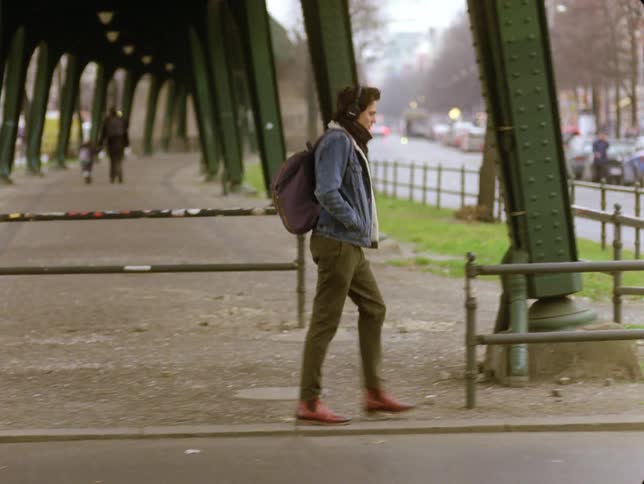 A young woman with a backpack strides under a bridge in the city streets
