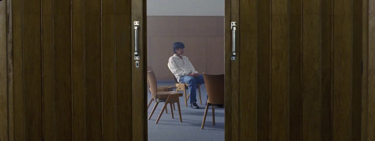 A man walks in a circle of chairs in an otherwise empty room and sits on one of the chairs