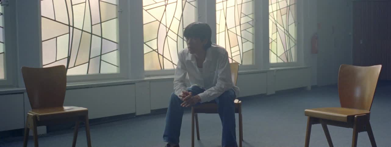 A man in a white shirt is sitting on a chair and there are several other chairs around him in an empty room and he is thoughtful