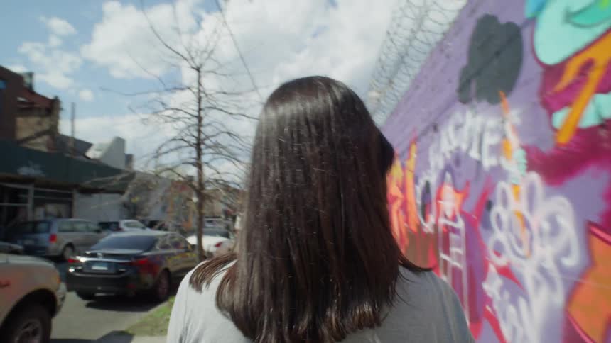 Perspective of a woman seen from behind walking on a sidewalk past a wall with graffiti