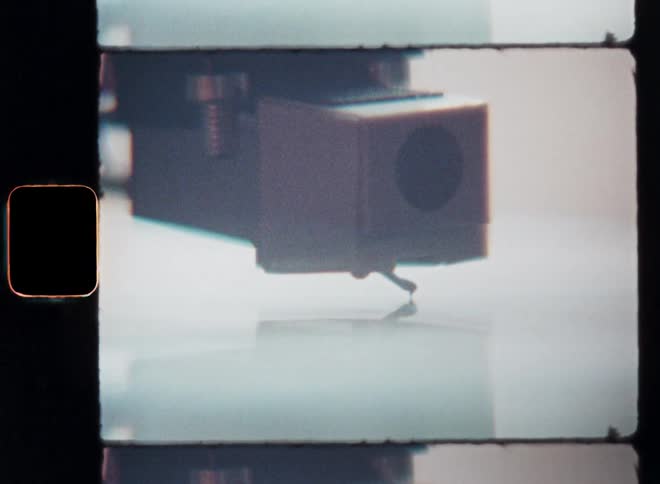 Super-8 macro closeup film footage of the player arm of a turntable lowering and the needle of the record player is moving down onto the vinyl record as the music starts playing or the song begins to sound on the old stereo system