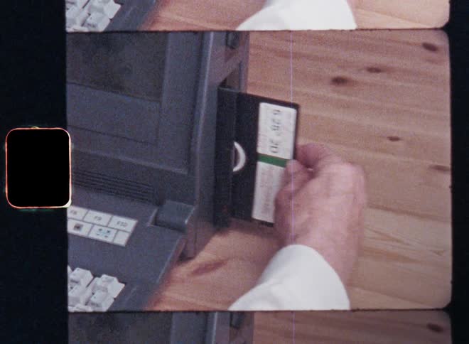 Retro Super 8 mm film footage with nostalgic vibe of a male person with white shirt putting a floppy disk into a vintage Osborne 3 computer laptop machine sitting at a wooden desk or table as symbol for technological progress and the digital revolution