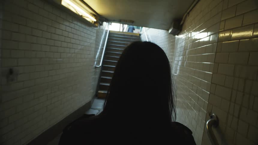 Perspective of a woman seen from behind climbing the stairs of a train station and emerging onto the sidewalk of a metropolis