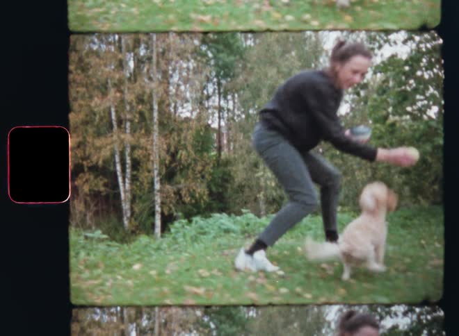 Young brunette woman walking or running in a garden or flat field with grass playing with a ball with her cute small poodle puppy that follows the pet owner holding the dog toy and a bowl of dog food in her hands on vintage 8mm film