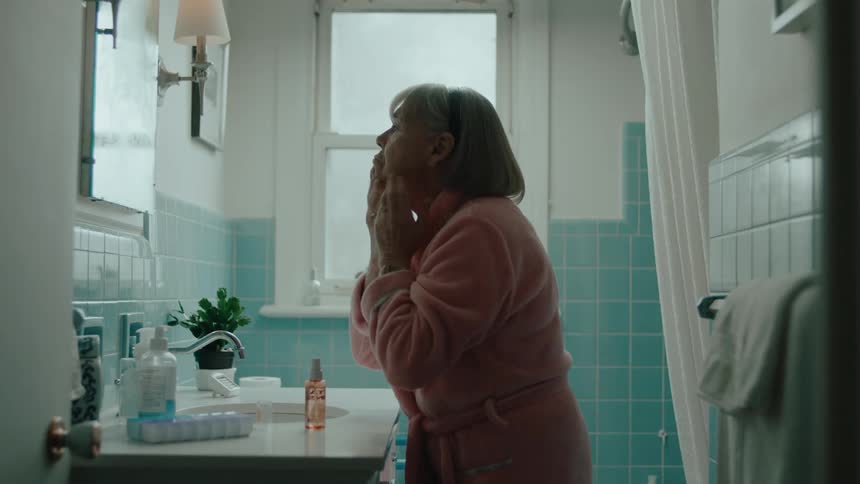 Older woman examines her face in bathroom mirror after spritzing her face