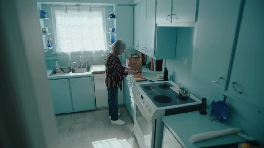 Elderly woman stands in kitchen while peeling and eating a carrot