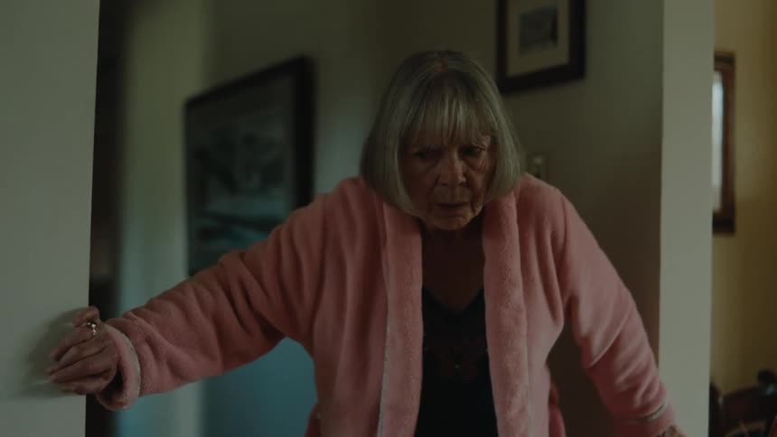 Elderly lady wearing a pink bathrobe walking around the house leaning on the walls with a weak facial expression