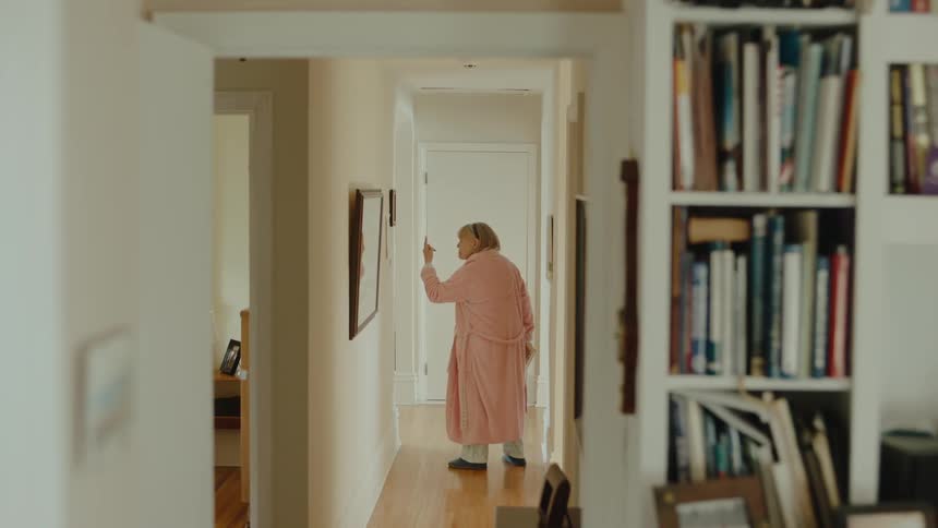 An elderly woman walks down a hallway and makes an obscene gesture to a picture on the wall