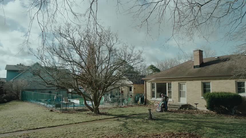 A man pushes a woman in a wheelchair across a yard