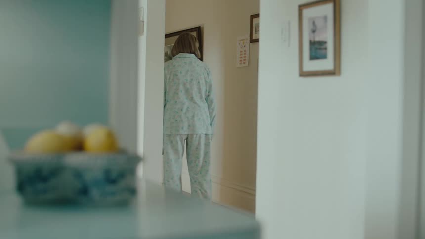 A woman limps down a hallway carrying a remote wearing pajamas and flips her middle finger at a piece of paper on the wall
