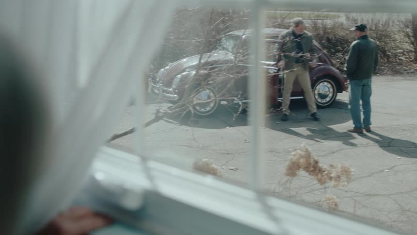 Elderly woman watching from the window two men talking next to a beetle
