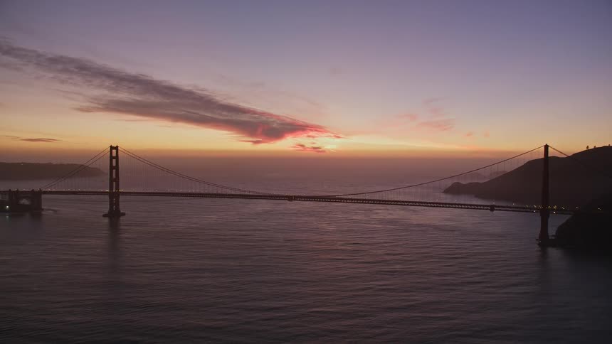Suspension bridge crossing ocean water stands silhouetted against the sunlight glowing on the red tinted horizon