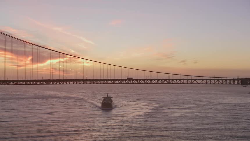 Aerial view of a boat passing under a suspension bridge at sunset