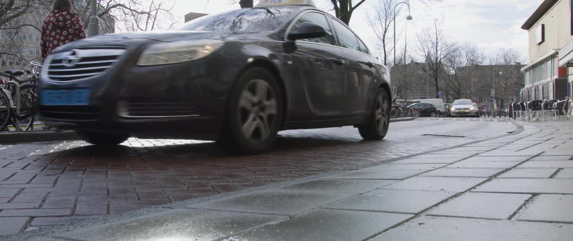 A taxi is driving on a cobblestone street beside a sidewalk with parked bicycles