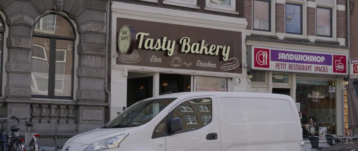 A street view featuring a bakery and sandwich shop storefronts with cars parked in front