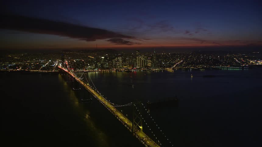 At night the san franciscooakland bay bridge is captured in an aerial view illuminated with yellow lights while red taillights and white headlights from vehicles create streaks of light as they traverse the bridge
