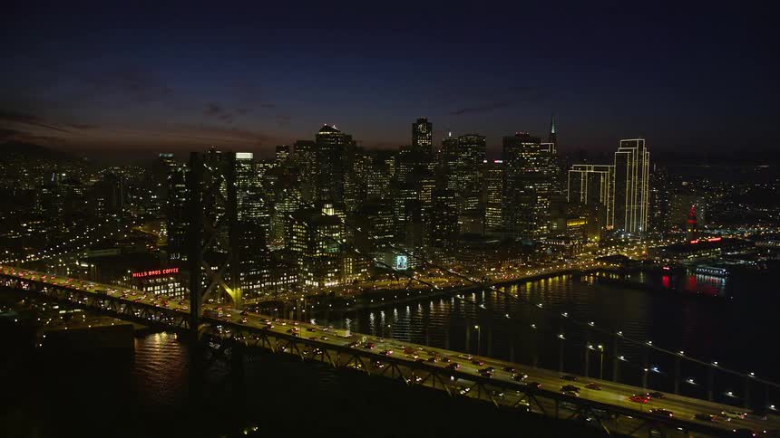 At night an aerial view of the san francisco bay area reveals twinkling lights from the bridge and the city contrasting against the dark expanse of the bay with the silhouette of alcatraz island visible in the distance