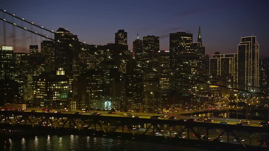 Aerial panorama in the foreground of an illuminated bridge at night with vehicle traffic and a seaside metropolis in the background