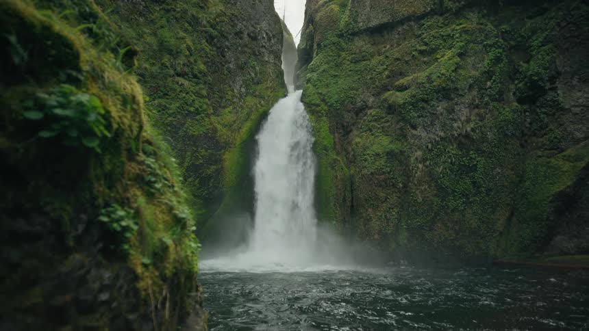 A waterfall flows between two mosscovered cliffs into a dark pool below
