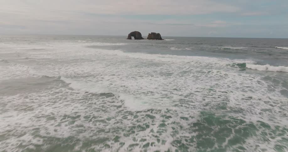 Beach with crashing waves and two isolated rocks in the middle of the sea on a blue sky day with clouds