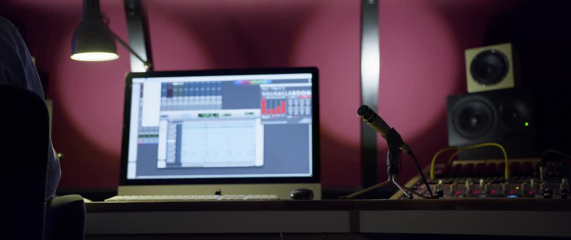 Person sits in a professional recording studio setup with a microphone computer monitor and audio mixer on a desk