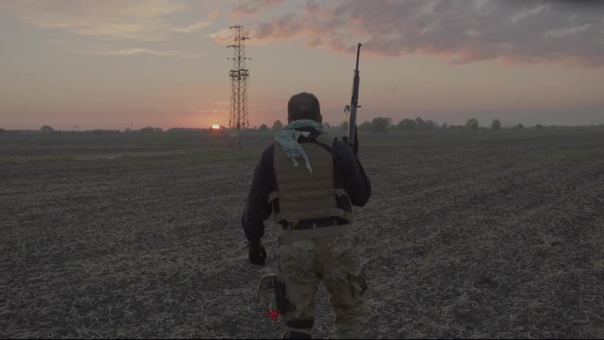 Man walking in a dry agriculture field holding a gun and wearing a vest
