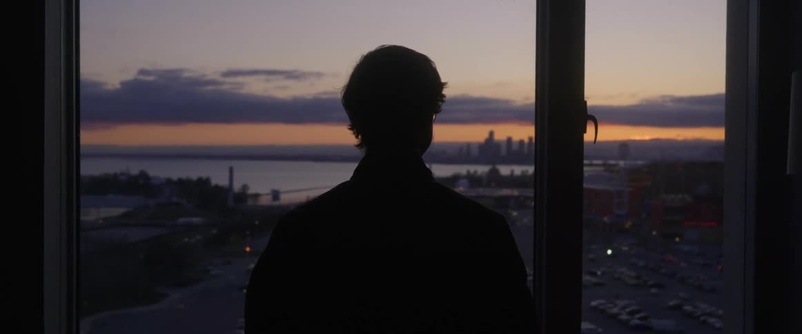 A man stands at a window and looks at the city below