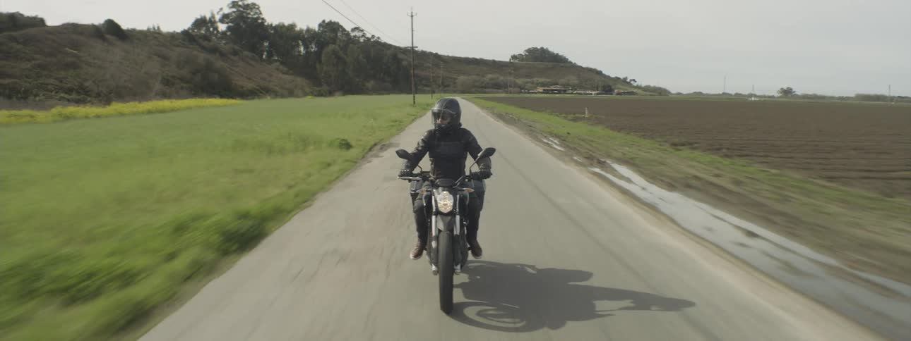 A motorcyclist in black clothes and helmet is driving on a street in a rural region
