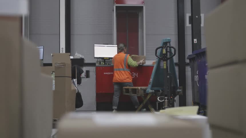 Woman with an orange vest inside a factory placing a box on a conveyor belt