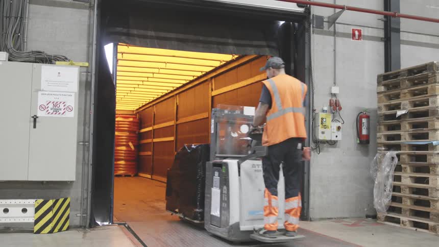 Man in an orange vest driving a cart that loads pallets inside a factory
