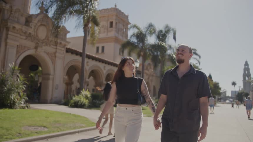 A man in a black shirt and pants and a woman wearing a black top a black bag and beige pants walk hand in hand at balboa park
