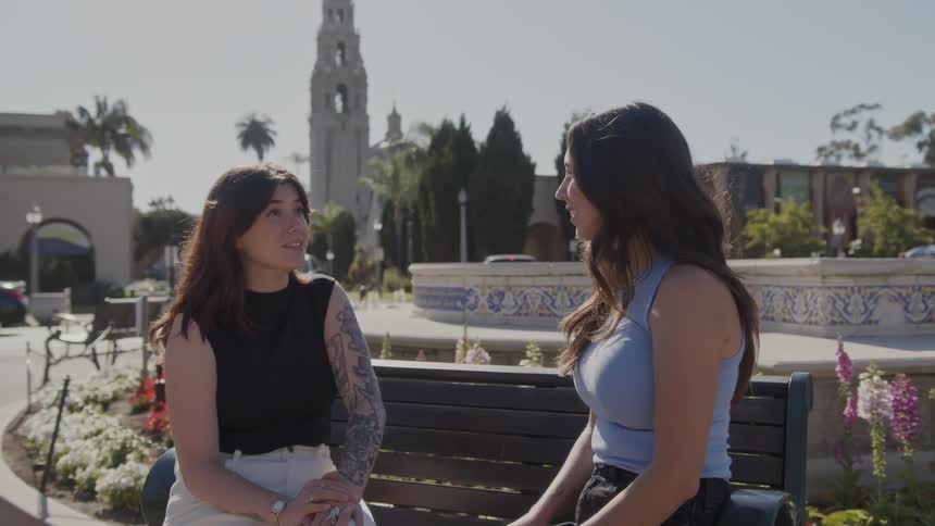 Two women sit on a bench outdoors and talk
