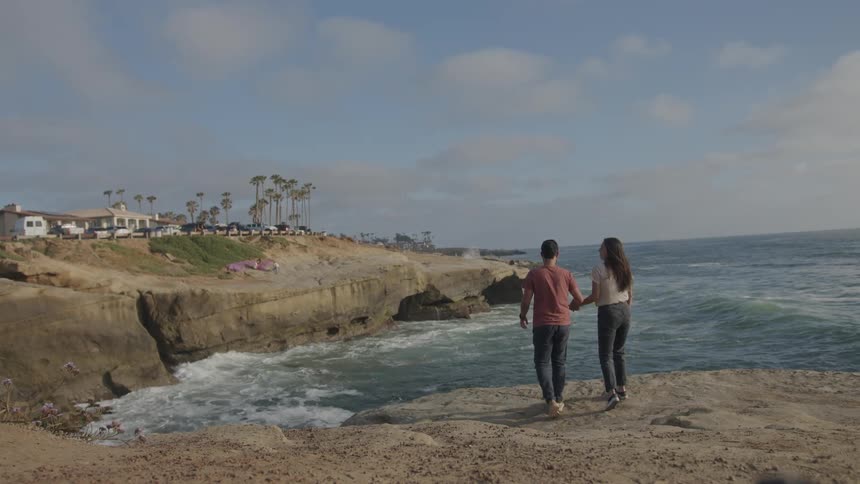 A couple stands on a rocky shoreline gazing at each other with the ocean and a flock of birds in the background capturing a serene and romantic moment by the sea