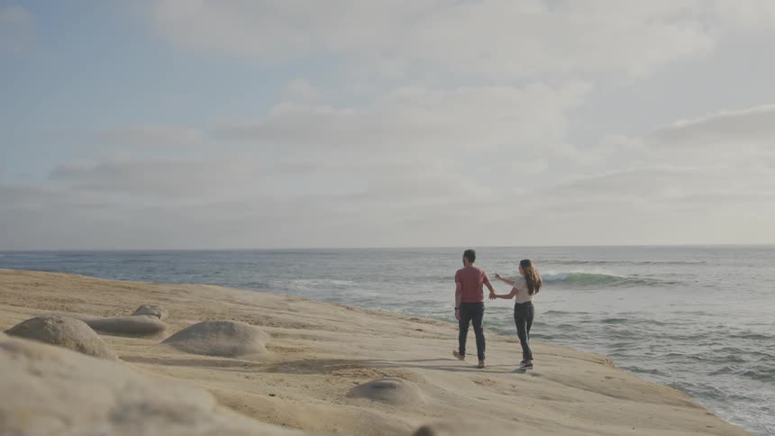 A young couple walk on a sandy beach holding hands next to the ocean