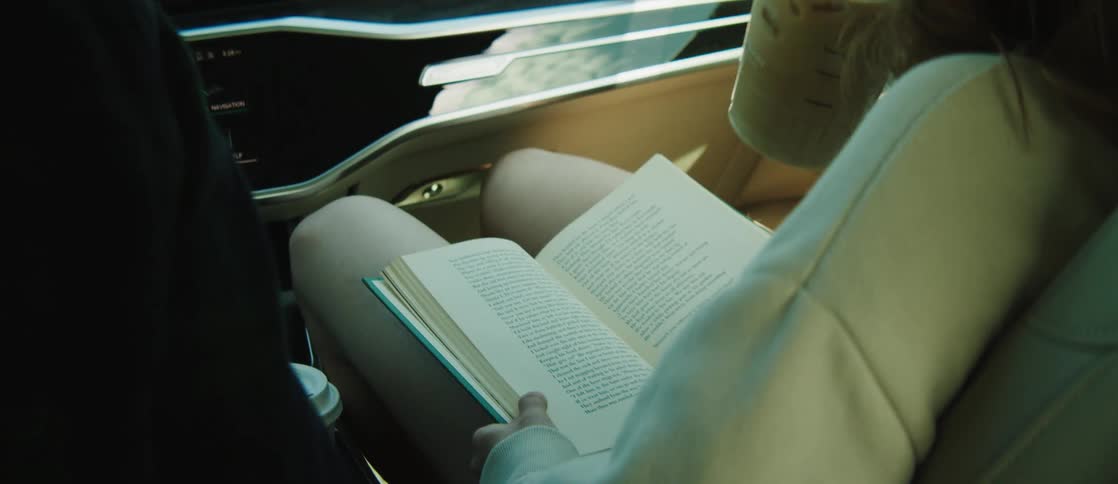 Woman sitting inside a car reading a book and taking a disposable cup with liquid and drinking it