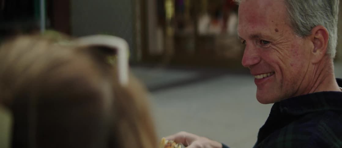 The middleaged man wearing a black shirt holds food in his hands and talks with a woman who is smiling and wearing sunglasses