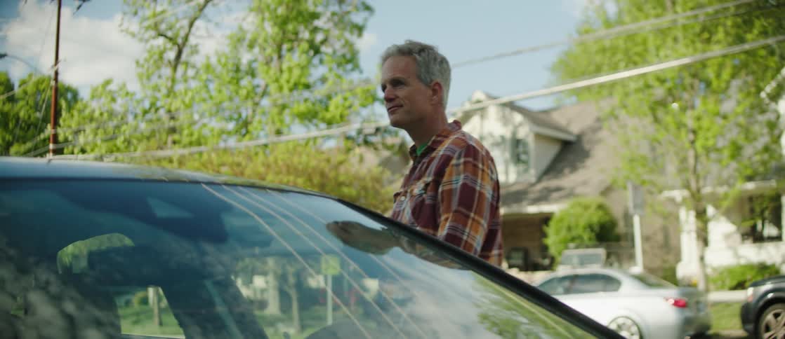 An individual in a plaid shirt and jeans stands beside a car in a residential area