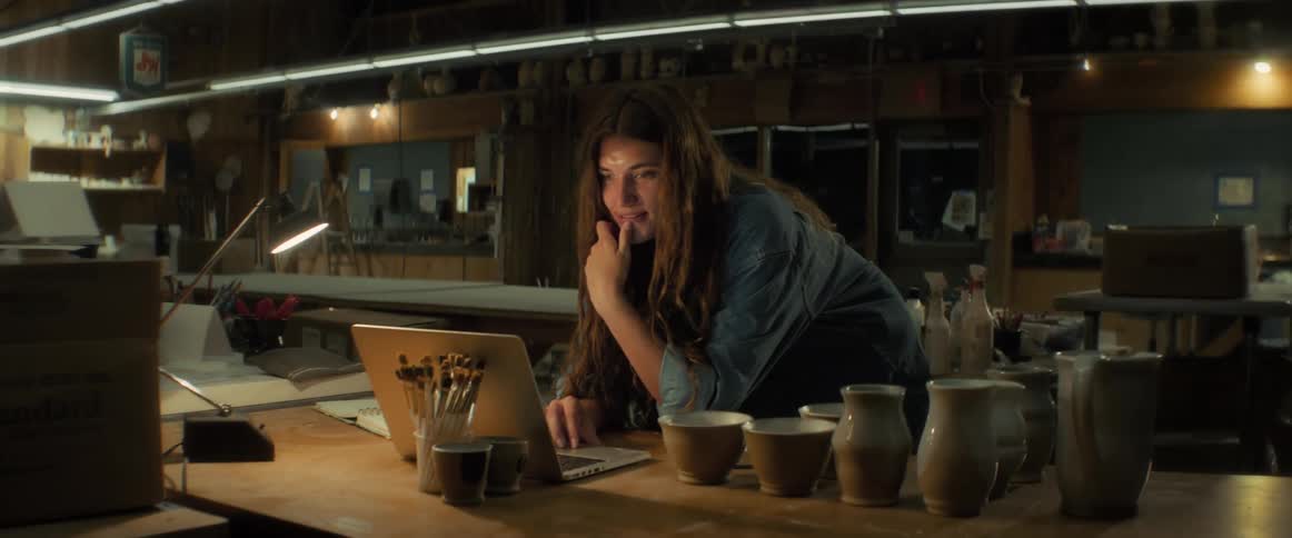 Happy woman with brown wavy hair and a blue shirt works on laptop in a workshop