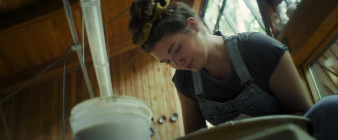 A woman with a headband and overalls is leaning over a container in a rustic indoor setting