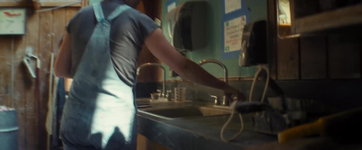 A woman with a bun hairstyle and a jumpsuit walks to the kitchen sink in a workshop to wash her hands