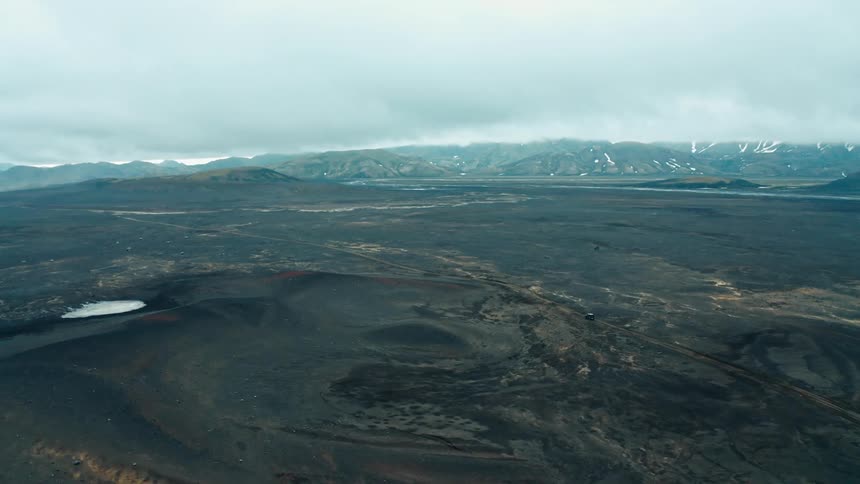 Aerial view of a vehicle driving through a volcanic landscape next to the mountains