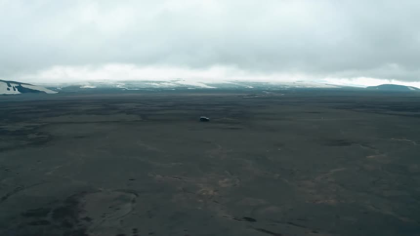 Aerial panorama of a vehicle driving through dry ground with snowy mountains around it on a cloudy day