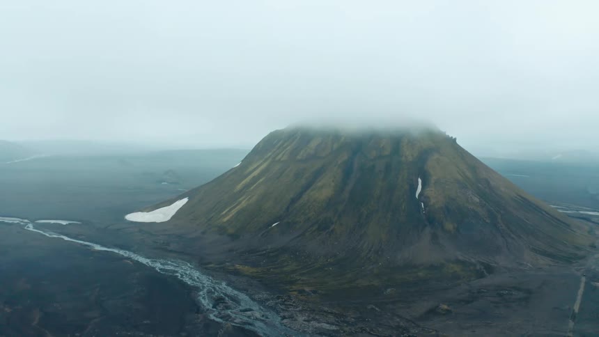 A large mountain covered in clouds next to a river and plain