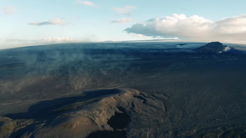 Aerial view of a vast desolate landscape featuring volcanic terrain with a distant view of a mountainous area and cloudcovered sky